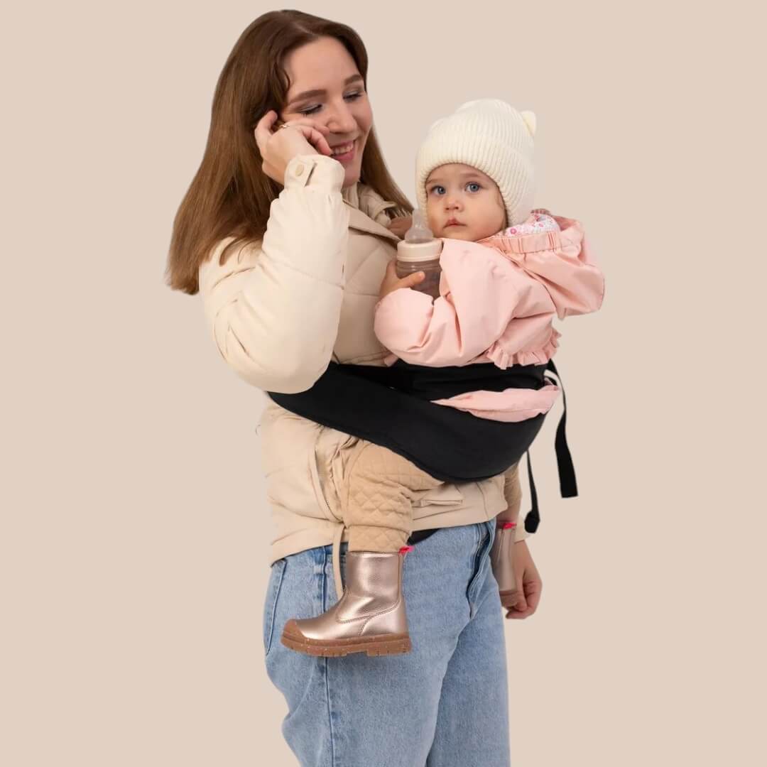 Maman portant son bébé sur la hanche avec un porte-bébé, tout en ayant les mains libres pour téléphoner.